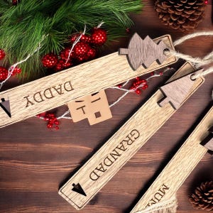 May include: Three wooden Christmas ornaments with a tree design and the words "Daddy", "Granddad", and "Mum" engraved on them. The ornaments are on a brown wooden surface.