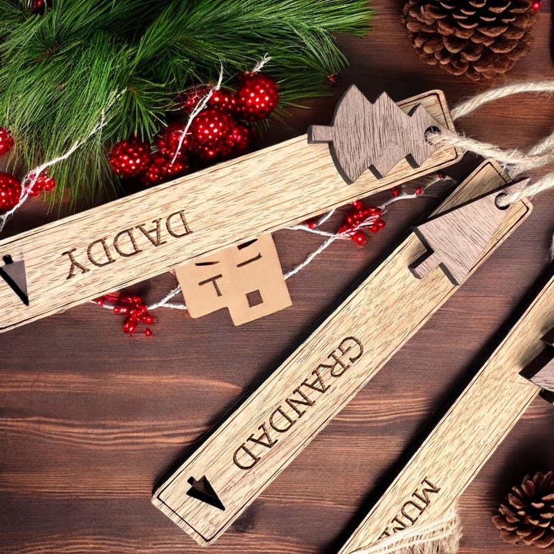 May include: Three wooden Christmas ornaments with a tree design and the words "Daddy", "Granddad", and "Mum" engraved on them. The ornaments are on a brown wooden surface.