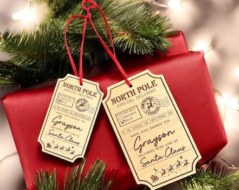 May include: Two wooden Christmas ornaments shaped like vintage mail tags. The tags are decorated with a North Pole theme and have the text "North Pole Special Delivery" and "Elf Use Only Naughty Nice". The tags are personalized with the name "Grayson" and the message "Delivered with love by Santa Claus".