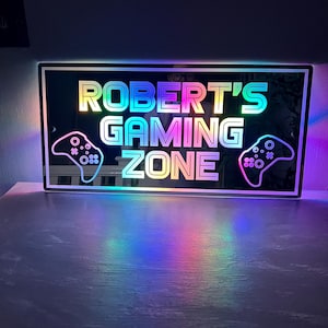 May include: A rectangular sign with the text "ROBERT'S GAMING ZONE" in rainbow-coloured letters. Two controller outlines flank the text. The sign is illuminated with a colourful glow, creating a vibrant and eye-catching display.