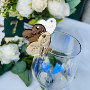 May include: Two wooden heart-shaped place cards with the names "Bethany" and "Olivia" written on them. The place cards are attached to a clear wine glass.