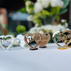 May include: Wooden heart-shaped wedding place cards with the names Olivia and Olivia written in cursive. The cards are on a white table with a bouquet of white flowers in the background.