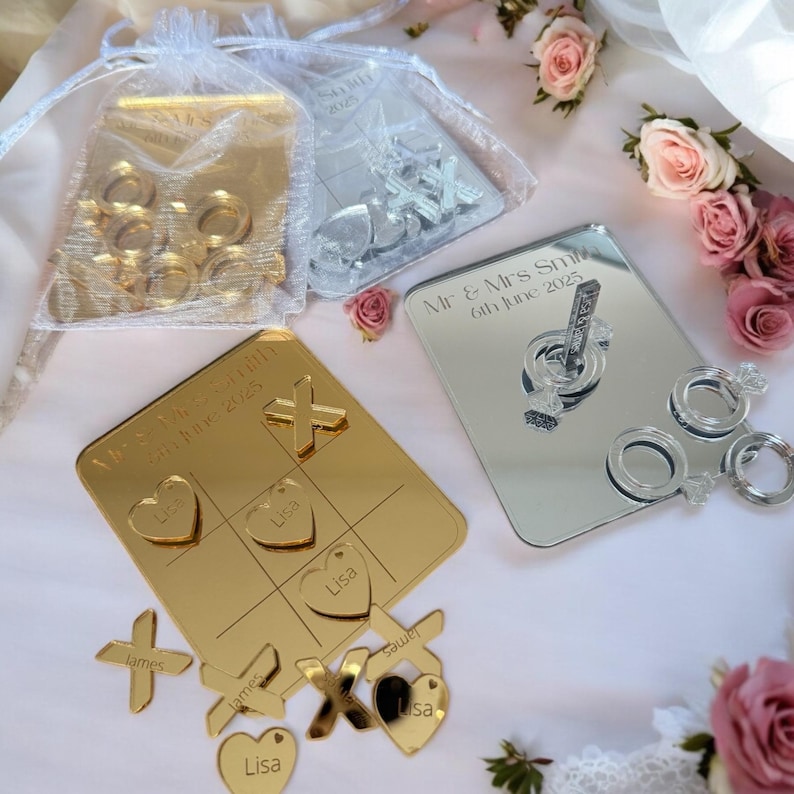 Puede incluir: Juego de tres en raya de espejo dorado y plateado con piezas en forma de coraz&oacute;n y X. El tablero de juego est&aacute; personalizado con los nombres "James" y "Lisa" y la fecha "6 de junio de 2025".