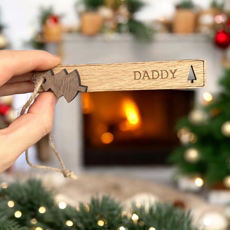 May include: Wooden Christmas ornament with a small tree cutout and the word "DADDY" engraved on it. The ornament is attached to a twine string.