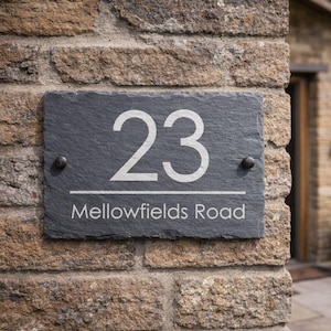 Op de afbeelding: Een rechthoekig leien huisnummerbord met het witte nummer "23" en de tekst "Mellowfields Road" eronder. Bevestigd aan een bakstenen muur met twee donkere schroeven.