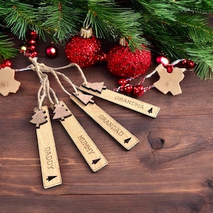 May include: Four wooden Christmas tree ornaments with the words "Daddy", "Mummy", "Grandad", and "Grandma" engraved on them. The ornaments are hanging from twine and are surrounded by red and green Christmas decorations.
