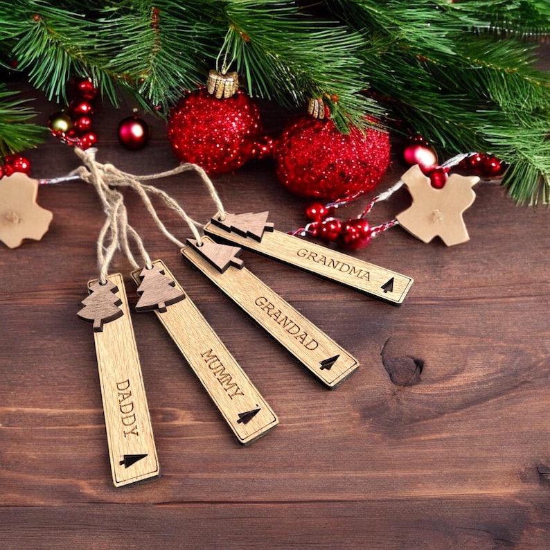 May include: Four wooden Christmas tree ornaments with the words "Daddy", "Mummy", "Grandad", and "Grandma" engraved on them. The ornaments are hanging from twine and are surrounded by red and green Christmas decorations.