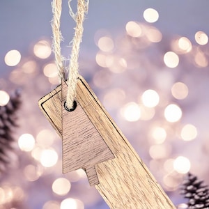 May include: A wooden Christmas tree ornament with a rustic brown finish. The ornament is hanging from a twine string and has a hole in the top for hanging. The ornament is in focus, while the background is blurred and features a bokeh effect of white lights.