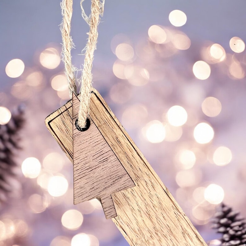 May include: A wooden Christmas tree ornament with a rustic brown finish. The ornament is hanging from a twine string and has a hole in the top for hanging. The ornament is in focus, while the background is blurred and features a bokeh effect of white lights.