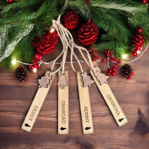 May include: Four wooden Christmas tree ornaments with the words "Daddy", "Grandad", "Mummy", and "Grandma" engraved on them. The ornaments are hanging from twine and are surrounded by pine branches, red berries, and pine cones.