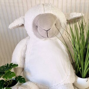 May include: A white plush lamb with a grey face and feet. The lamb is sitting on a white fluffy surface.