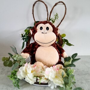 May include: A brown plush monkey toy sitting in a wreath of greenery and white flowers. The monkey has a brown face and a light brown body.