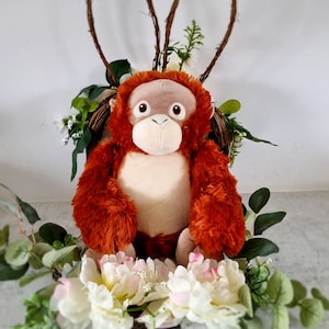 May include: A plush orange orangutan monkey toy sitting in a wicker basket decorated with white and green flowers.