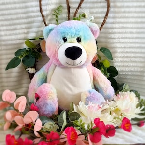 May include: A rainbow tie-dye plush teddy bear with a white belly and black nose, sitting in a bed of pink and white flowers.