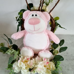 May include: Pink plush teddy bear with a white tummy, sitting in a bed of white and pink flowers. The bear has a pink nose and black eyes.