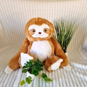 May include: A plush brown and white sloth toy with a friendly face, sitting on a white fluffy surface with a green potted plant.