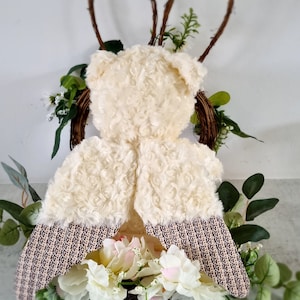 May include: A white teddy bear with wings made of white fabric with a brown and white patterned trim. The wings are spread out and the bear is sitting on a bed of white flowers.
