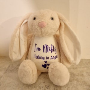 May include: A beige plush bunny with long floppy ears. The bunny has a white patch on its chest with purple text that reads "I'm Mifty I belong to Angel" and three purple hearts.