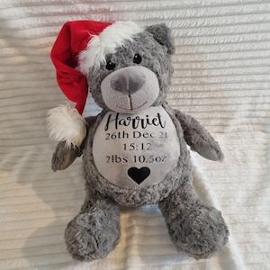May include: A gray plush teddy bear wearing a red Santa hat with white trim. The bear has a white patch on its chest with the text "Harriet 26th Dec 21 15:12 7lbs 10.5oz" and a black heart.
