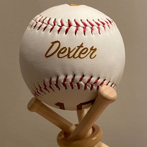 May include: White baseball with red stitching and the name "Dexter" written in gold lettering. The baseball is displayed on a wooden stand with two crossed bats.