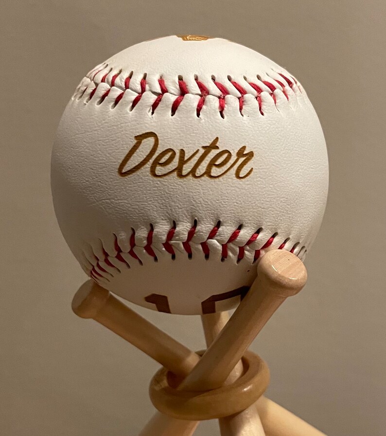 May include: White baseball with red stitching and the name "Dexter" written in gold lettering. The baseball is displayed on a wooden stand with two crossed bats.