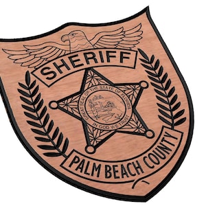 Puede incluir: Insignia de sheriff de madera con el texto "SHERIFF" y "PALM BEACH COUNTY". La insignia presenta un águila, una estrella con las palabras "GREAT SEAL OF THE STATE OF FLORIDA" y el lema "IN GOD WE TRUST".