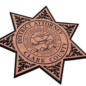 May include: A wooden star-shaped plaque with a black outline and a seal in the centre. The seal reads "The Great Seal of the State of Nevada" and "All for Our County". The plaque also reads "District Attorney Clark County".