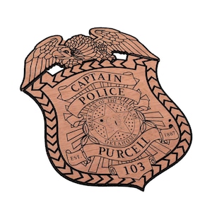 May include: A wooden police badge with a brown background and black outline. The badge features an eagle, the words "Captain Police", the seal of the state of Oklahoma, the text "Purcell" and the number "103".