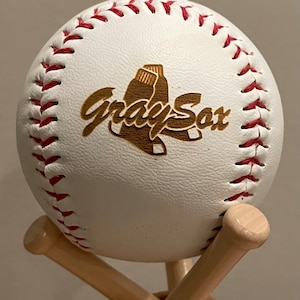 May include: A white baseball with red stitching and a brown wooden logo that says "Gray Sox" with a baseball glove graphic. The baseball is on a wooden baseball bat stand.