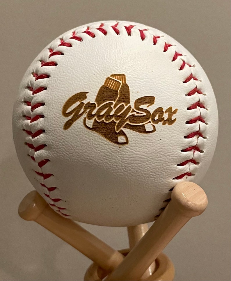 May include: A white baseball with red stitching and a brown wooden logo that says "Gray Sox" with a baseball glove graphic. The baseball is on a wooden baseball bat stand.