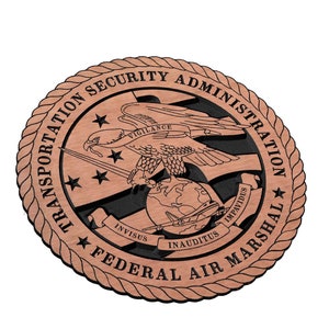 Puede incluir: Una placa redonda de madera con un borde de cuerda. La placa presenta el sello de la Administración de Seguridad del Transporte, Federal Air Marshal. El sello representa un águila con las alas extendidas, una espada, un globo terráqueo y tres estrellas. El texto "Transportation Security Administration" y "Federal Air Marshal" está inscrito alrededor del sello. Las frases latinas "Invisus, Inauditus, Impavidus" están inscritas en una pancarta debajo del globo.