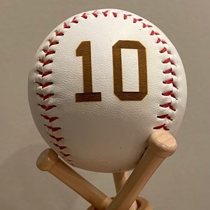 May include: A white baseball with red stitching and the number 10 in gold on it. The baseball is sitting on a wooden baseball bat stand.