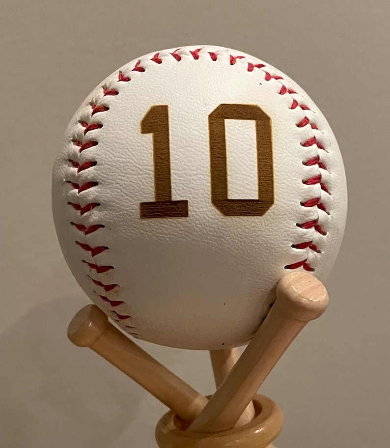 May include: A white baseball with red stitching and the number 10 in gold on it. The baseball is sitting on a wooden baseball bat stand.