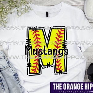 May include: White t-shirt with a black outline of the letter M. The letter is filled with a yellow softball design with red stitching. The word "Mustangs" is written in black inside the letter. The t-shirt is on a wooden surface.