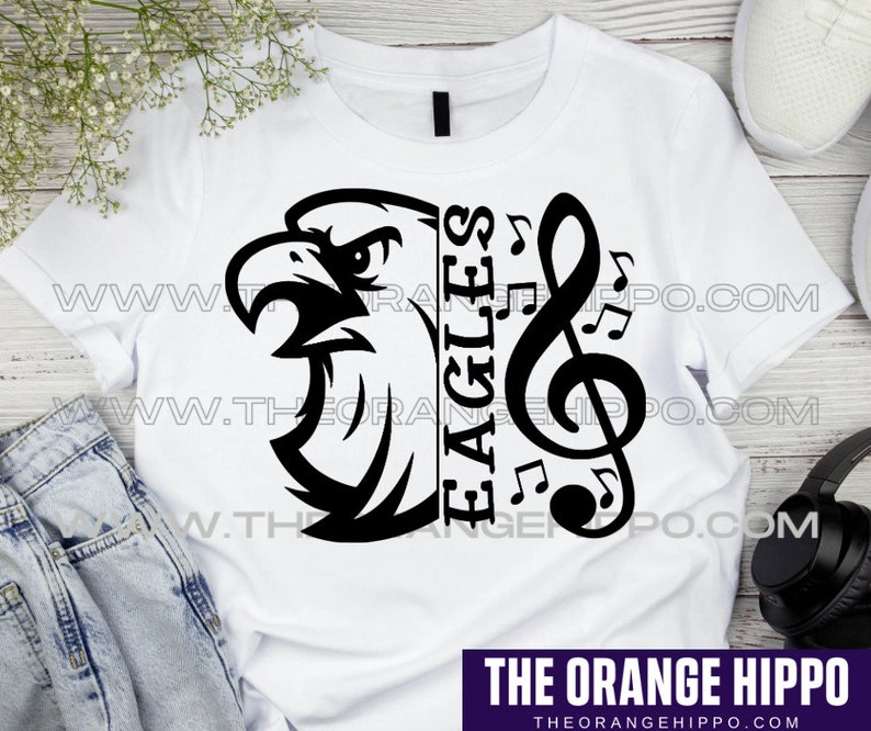 May include: White t-shirt featuring a black graphic of an eagle's head, musical notes, and the word "EAGLES". The design includes a treble clef and musical notes. The text "THE ORANGE HIPPO" is at the bottom.