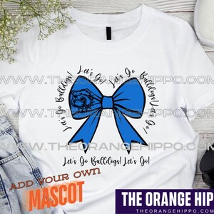 May include: White t-shirt with a blue bow and the text "Let's Go Bulldogs! Let's Go!"  The text is in a black circle with a bulldog head graphic. The text "Add Your Own Mascot" is at the bottom of the shirt.