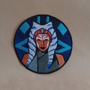 Könnte beinhalten: Gestickter Aufnäher mit einer blauen und weißen Figur mit orangefarbener Haut und blauem Hintergrund. Die Figur trägt weiße Stirnbänder und einen blauen und weißen Kopfschmuck. Der Hintergrund ist ein dunkelblauer Kreis mit einem hellblauen und weißen Design.