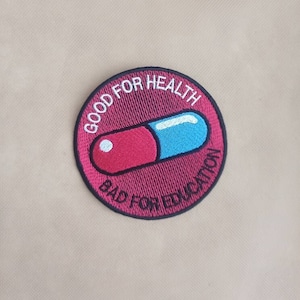 May include: Embroidered patch featuring a red and blue capsule design within a circular frame. The patch has the text "GOOD FOR HEALTH" and "BAD FOR EDUCATION" in white lettering, set against a maroon background.