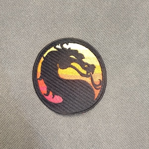 May include: A round, black embroidered patch featuring a dragon silhouette in a yellow, orange, and red gradient. The patch has a black border and is set against a gray fabric background.