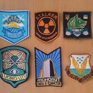 Könnte beinhalten: Sechs gestickte Aufnäher mit verschiedenen Designs. Einer zeigt eine Sonne und Vögel, ein anderer ein Strahlungssymbol und ein dritter ein wolfsähnliches Wesen. Weitere Aufnäher zeigen Atomsymbole, ein Gebäude und ein geflügeltes Emblem. Jeder Aufnäher hat Text.