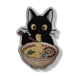Könnte beinhalten: Ein gestickter Aufnäher mit einer schwarzen Katze mit großen gelben Augen, die braune Stäbchen hält. Die Katze isst gelbe Nudeln aus einer beigen Schüssel, die eine grüne Garnierung und einen rosa-weißen Fischkuchen enthält.