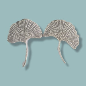 May include: Two embroidered, silver ginkgo leaf appliques. Each leaf features detailed veining and a long, slender stem. The appliques are set against a light blue background, creating a soft, natural aesthetic.