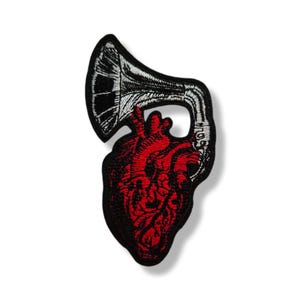 Peut inclure: Écusson brodé représentant un cœur anatomique rouge combiné à un gramophone noir et blanc. Le patch a une bordure noire et est conçu pour être appliqué sur du tissu. Le design est détaillé et artistique.