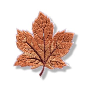 Può includere: Una toppa ricamata a forma di foglia d'acero, caratterizzata da sfumature di rame, bronzo e filo marrone chiaro. Il design include cuciture dettagliate per rappresentare le venature della foglia, con un bordo bianco che delinea la forma.