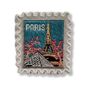 May include: Embroidered patch depicting the Eiffel Tower and the word "PARIS" on a teal background. The patch has a white, scalloped border and includes pink and gold floral accents.