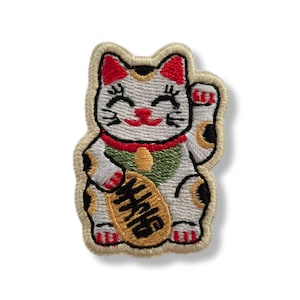 Puede incluir: Un parche bordado de un gato de la suerte blanco con manchas negras y doradas, orejas rojas y un collar verde. El gato tiene una cara sonriente y sostiene una moneda ovalada dorada con caracteres japoneses negros en su pata derecha.