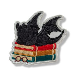 Puede incluir: Un parche bordado que muestra un pequeño dragón negro durmiendo sobre una pila de tres libros. Los libros son de color rojo, amarillo y verde azulado. Un par de gafas redondas de color rojo oscuro descansa sobre el libro inferior. Borde blanco.