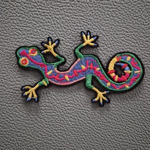 May include: An embroidered patch in the shape of a lizard with a curled tail. The lizard features a blue body with a pink stripe, yellow accents on the feet and back, and a multi-colored tail with pink, yellow, and blue segments outlined in black.