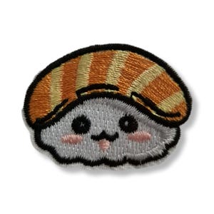 Könnte beinhalten: Ein gestickter Aufnäher, der ein Sushi-Nigiri-Cartoon-Motiv mit einer weißen Reisbasis und einem gestreiften orange-gelben Fisch-Topping zeigt. Die Figur hat schwarze Augen, einen kleinen schwarzen Mund und hellrosa Wangen.