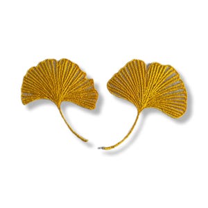 May include: Two gold-coloured ginkgo leaf-shaped appliques. Each leaf has a detailed, textured design with a stem. The appliques are set against a white background, creating a striking contrast. These could be used for crafting or embellishments.